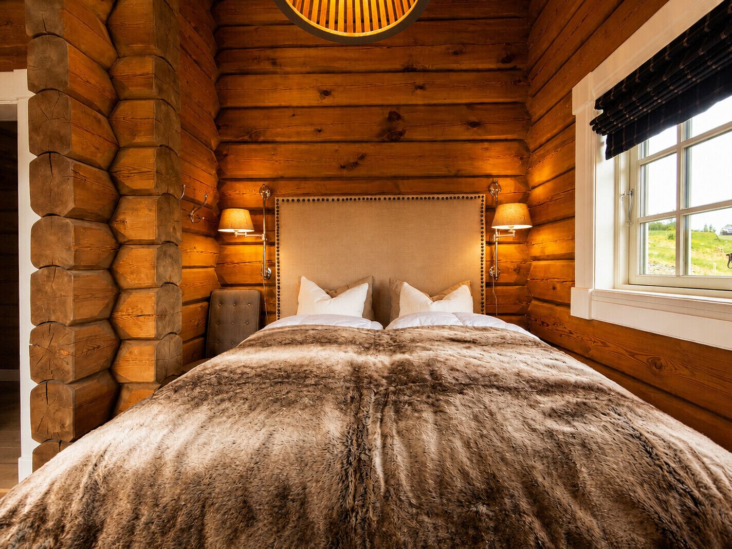 A log cabin bedroom with a wooden headboard, two bedside lamps, and a large fur-covered bed.