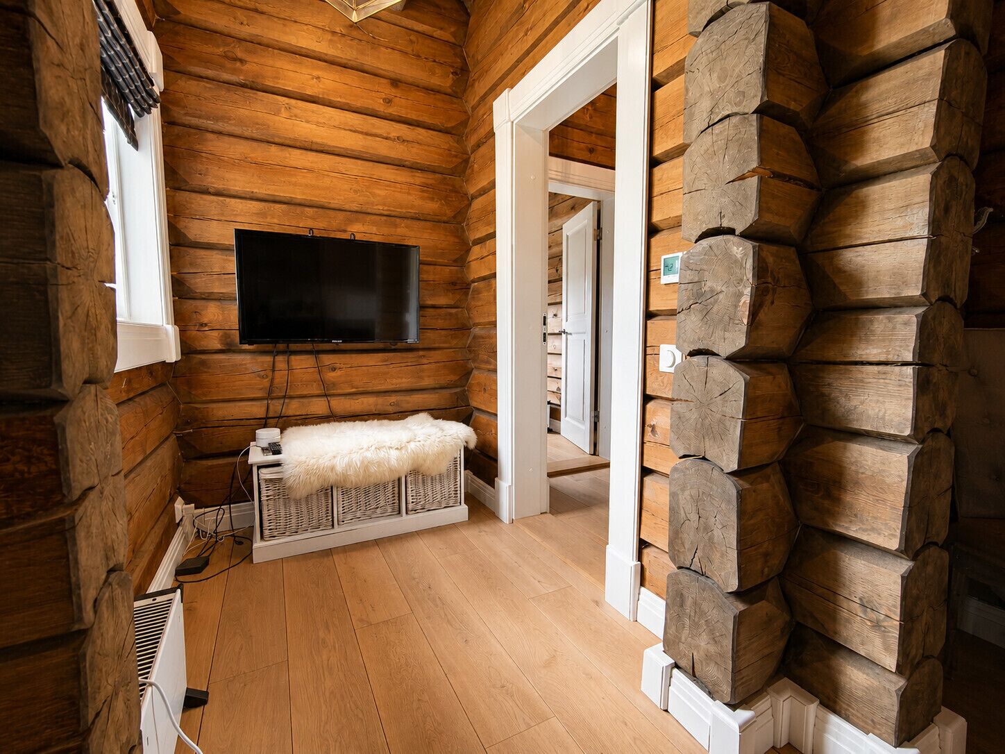 Interieur van een blokhut, met een televisie aan de wand, een witte bank met een bont plaid en een houten trap.