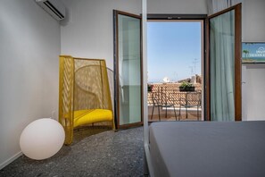 Habitación de hotel con cama, una silla amarilla, un objeto redondo blanco y un balcón con mobiliario exterior.