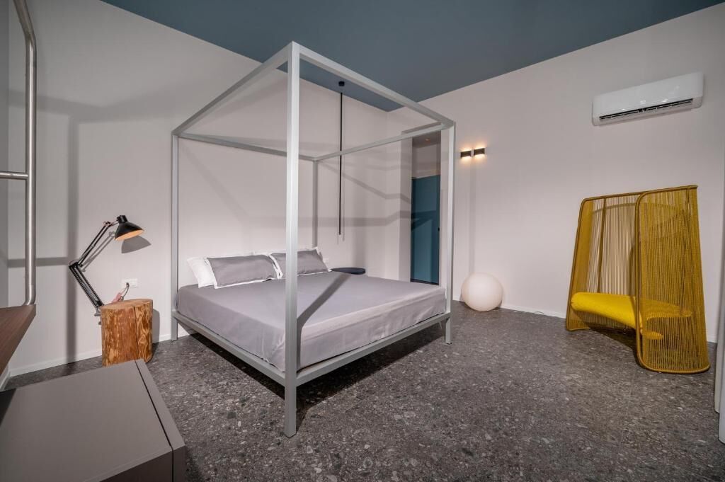 Ein modernes Schlafzimmer mit einem Himmelbett, einer Nachttischlampe, einem kleinen Holfnachtkästchen, einem gelben Sessel und einer weißen, kugelförmigen Dekoration am Boden.