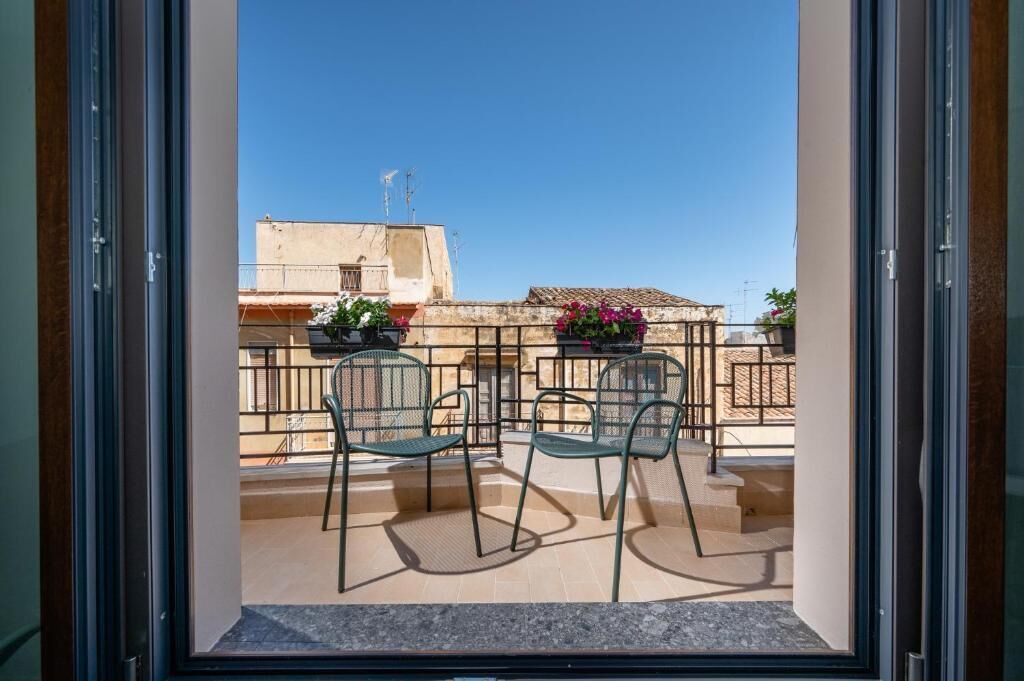 An open balcony with two chairs and a table, overlooking a residential area with buildings and a clear sky.