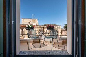 Un balcón con dos sillas y una mesa, con vista a un edificio de techo de tejas y cielo despejado.