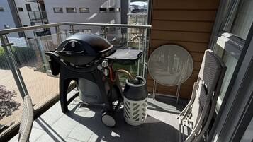 A balcony with a grill, a table, chairs, and a water bottle.
