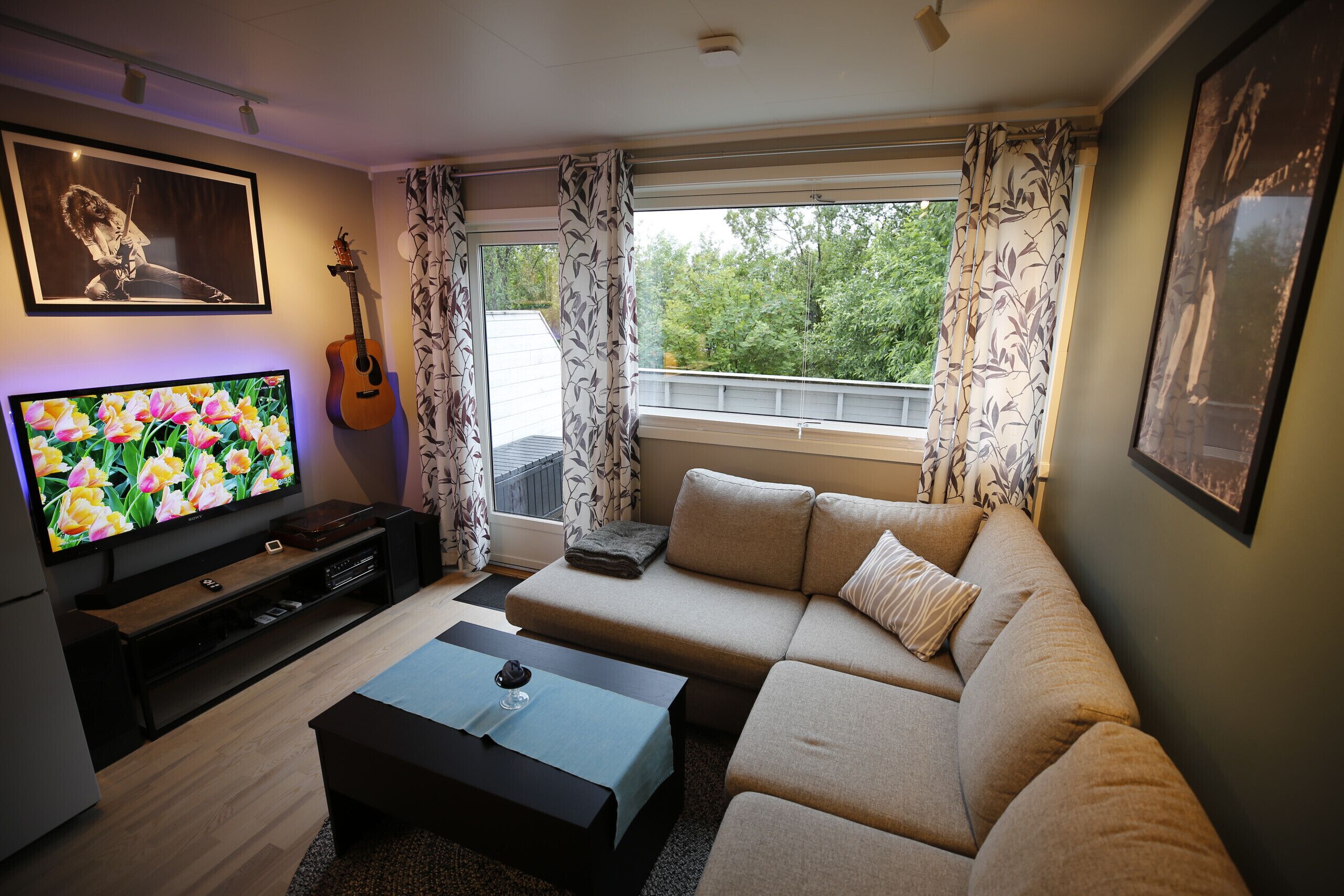 A living room with a sofa, a coffee table, a television, a guitar, and a balcony with curtains.