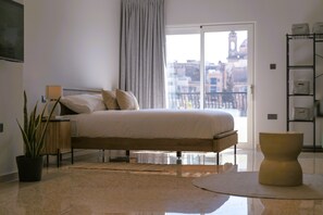 A bedroom with a bed, a nightstand, a potted plant, a chair, and a view of buildings through a window. - SIX • Birgu - 3-bed 3-bath luxury Marina Residence - Valletta ferry (Birgu)