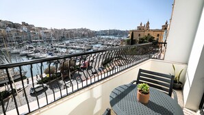Un balcón con mesa y sillas, con vista a un puerto deportivo con barcos y un paisaje urbano al fondo.