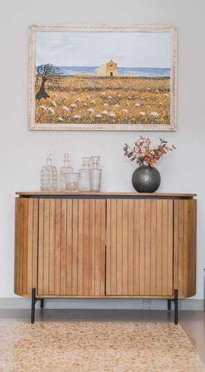 Un meuble en bois contenant des bouteilles en verre et surmonté d’un vase, ainsi qu’un tableau représentant un champ et un bâtiment accroché au mur au-dessus.