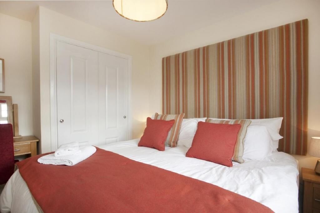 A hotel room with a bed, pillows, a red blanket, a striped headboard, a white door, and a nightstand with a lamp.