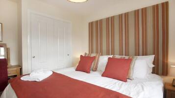 A hotel room with a bed, pillows, a red blanket, a striped headboard, a white door, and a nightstand with a lamp.