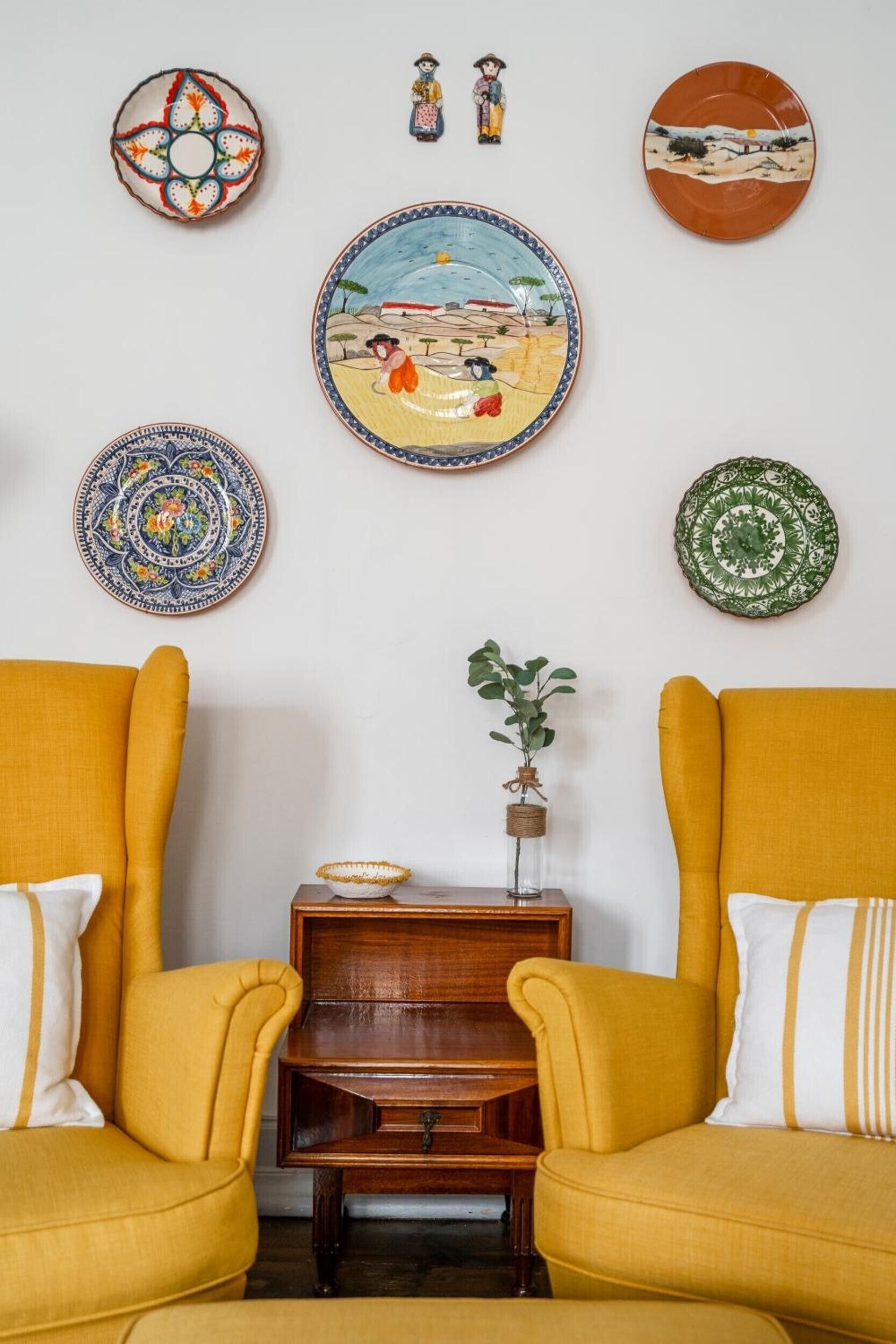 A room with two yellow armchairs, a small wooden side table, and a wall decorated with various plates and a small figurine.