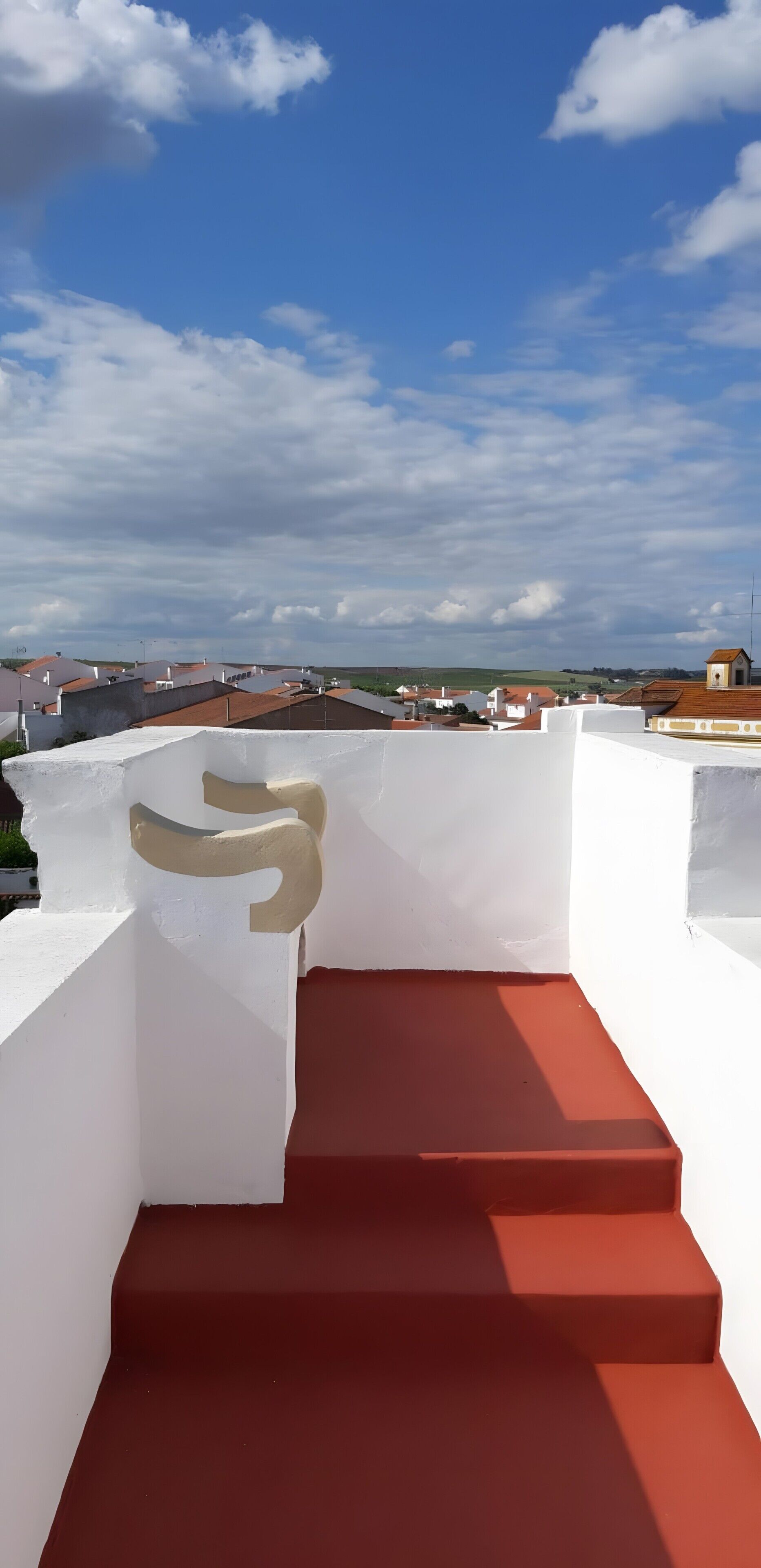 Una escalera roja que conduce a una pared blanca con una letra 'S' color beige, con vistas a una zona residencial con casas y un cielo despejado.
