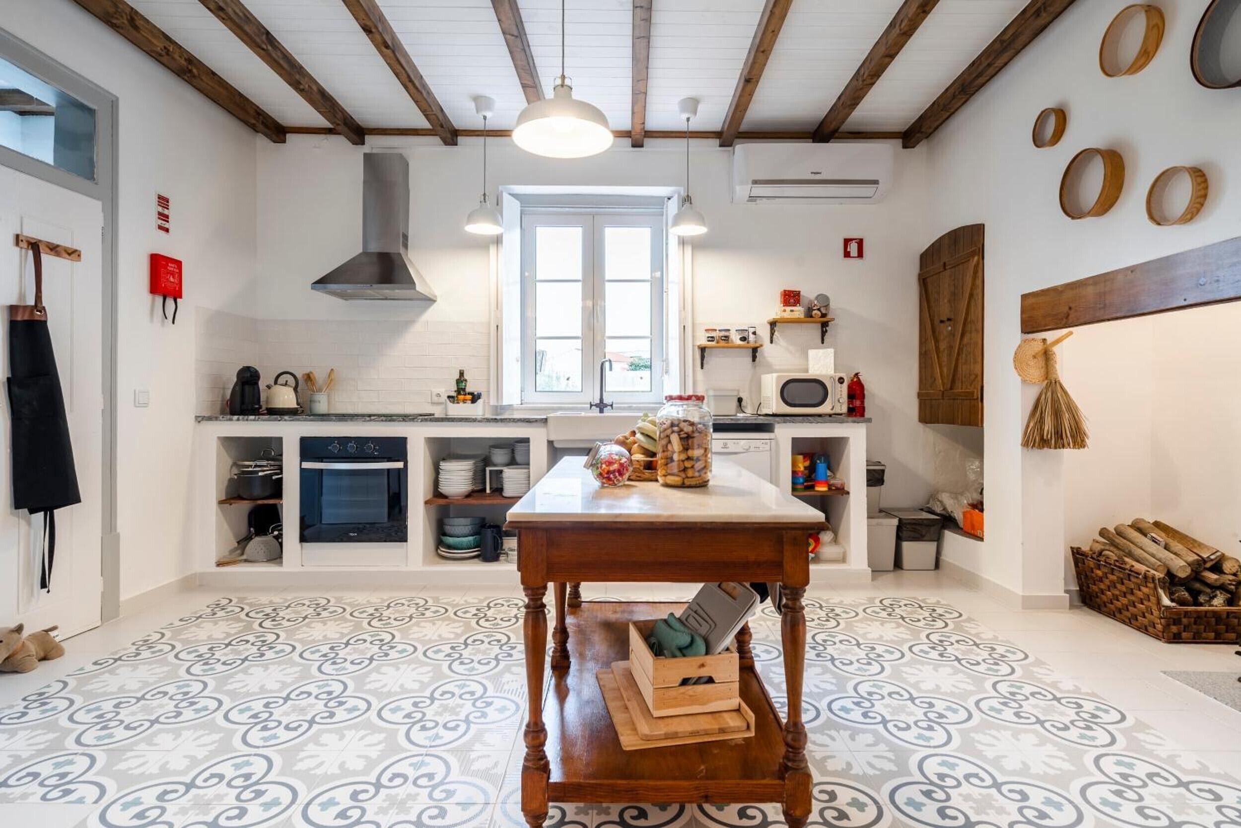 A kitchen with a central wooden table, white tiled floor, and a window above the sink.