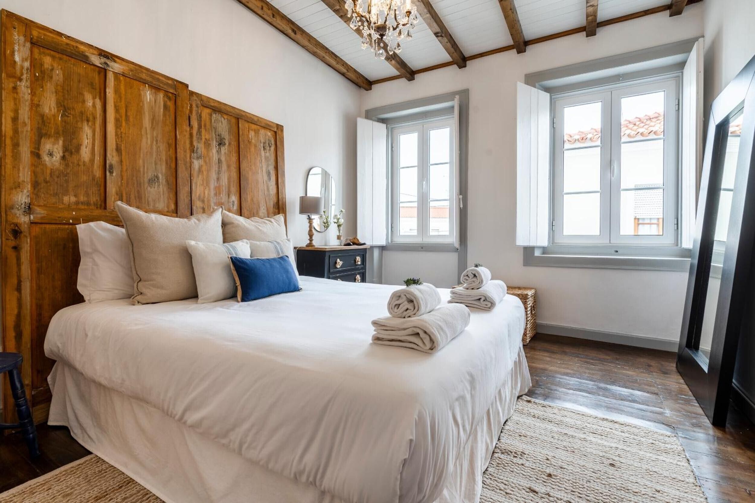 Ein Schlafzimmer mit einem großen Bett, einem Holz-Kopfteil, zwei Fenstern, einer Kronleuchter und einem kleinen Nachttisch mit Lampe.