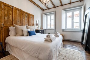Un dormitorio con una cama grande, cabecera de madera, dos ventanas, una lámpara de araña y una mesita de noche con lámpara.