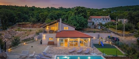A two-story house with a swimming pool, outdoor seating, and a view of the surrounding landscape.