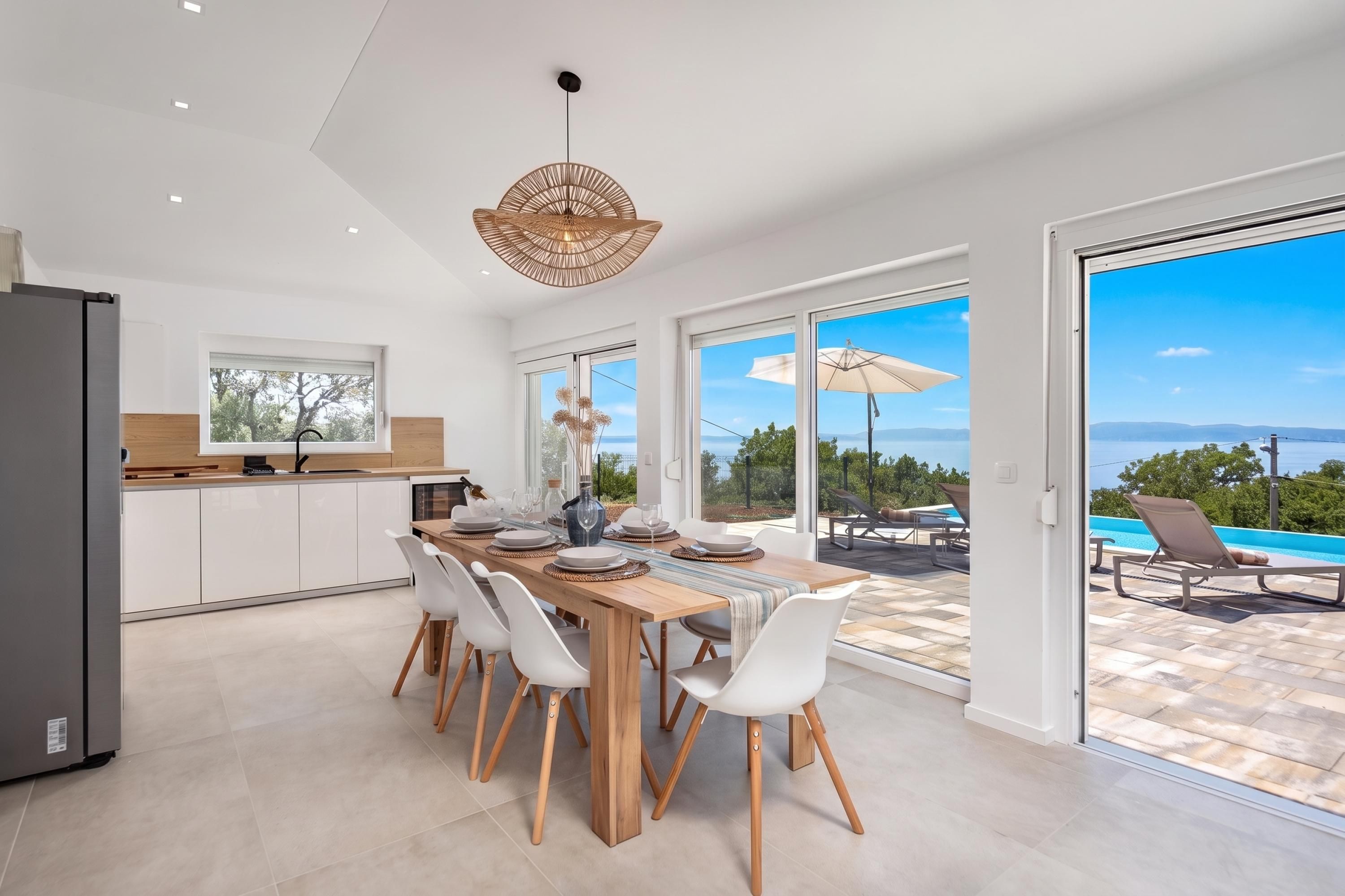 Un espace repas moderne avec une table en bois, des chaises blanches et une vue sur la mer grâce à de grandes fenêtres.