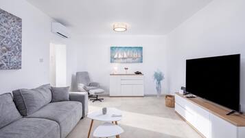 A modern living room with a grey sofa, a small white table, a TV on a wooden stand, and a painting of sailboats on the wall.