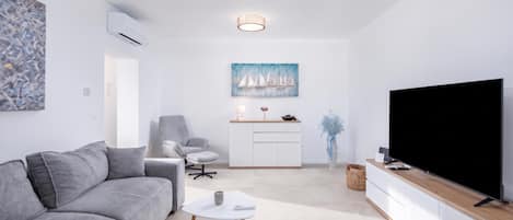 Ein modernes Wohnzimmer mit einem grauen Sofa, einem kleinen weißen Tisch, einem Fernseher auf einem Holzgestell und einem Gemälde von Segelbooten an der Wand.