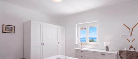 A minimalist bedroom with a large bed, a wardrobe, a window with a view, and a bedside table with a lamp.