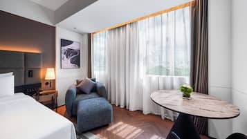 A modern hotel room with a bed, a sofa, a small table, and a view of the city through large windows.