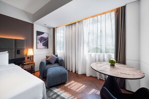 Habitación de hotel moderna con cama, sofá, una mesita y vistas a la ciudad a través de grandes ventanales.
