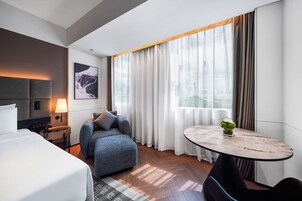 Una camera d'albergo moderna con un letto, una poltrona, un tavolino e vista sulla città attraverso ampie finestre.