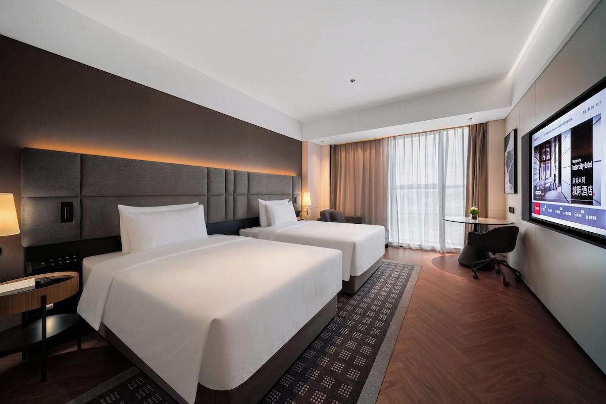 A hotel room with two beds, a desk, a chair, a TV, and a window with curtains.