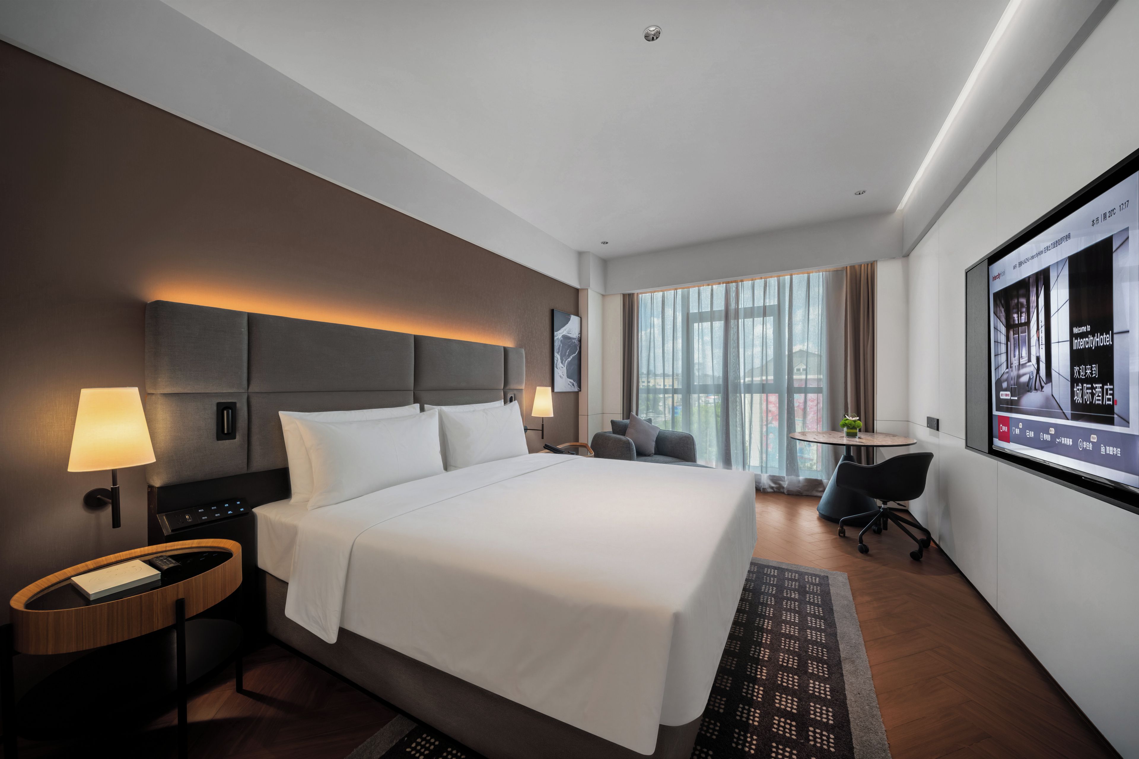 A modern hotel room with a large bed, bedside lamps, a desk, and a television screen displaying a welcome message.