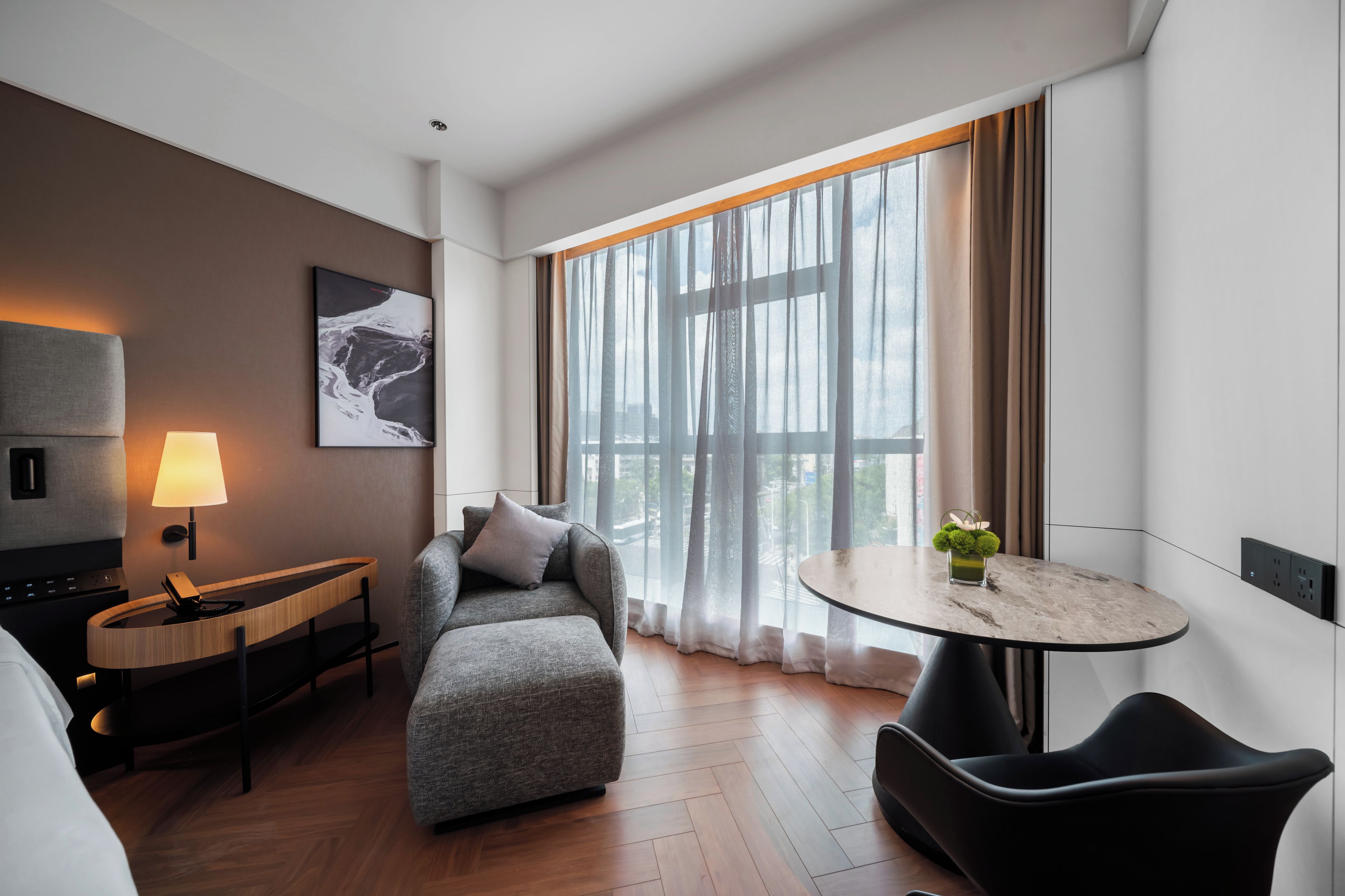 A modern hotel room with a sofa, a small table with a plant, a lamp, and a painting on the wall.