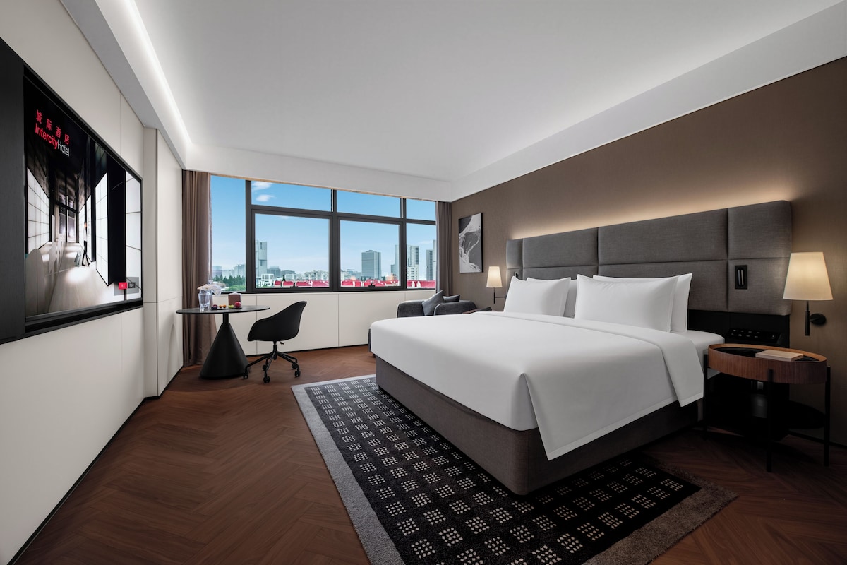 A modern hotel room with a large bed, a desk, and a view of the city.