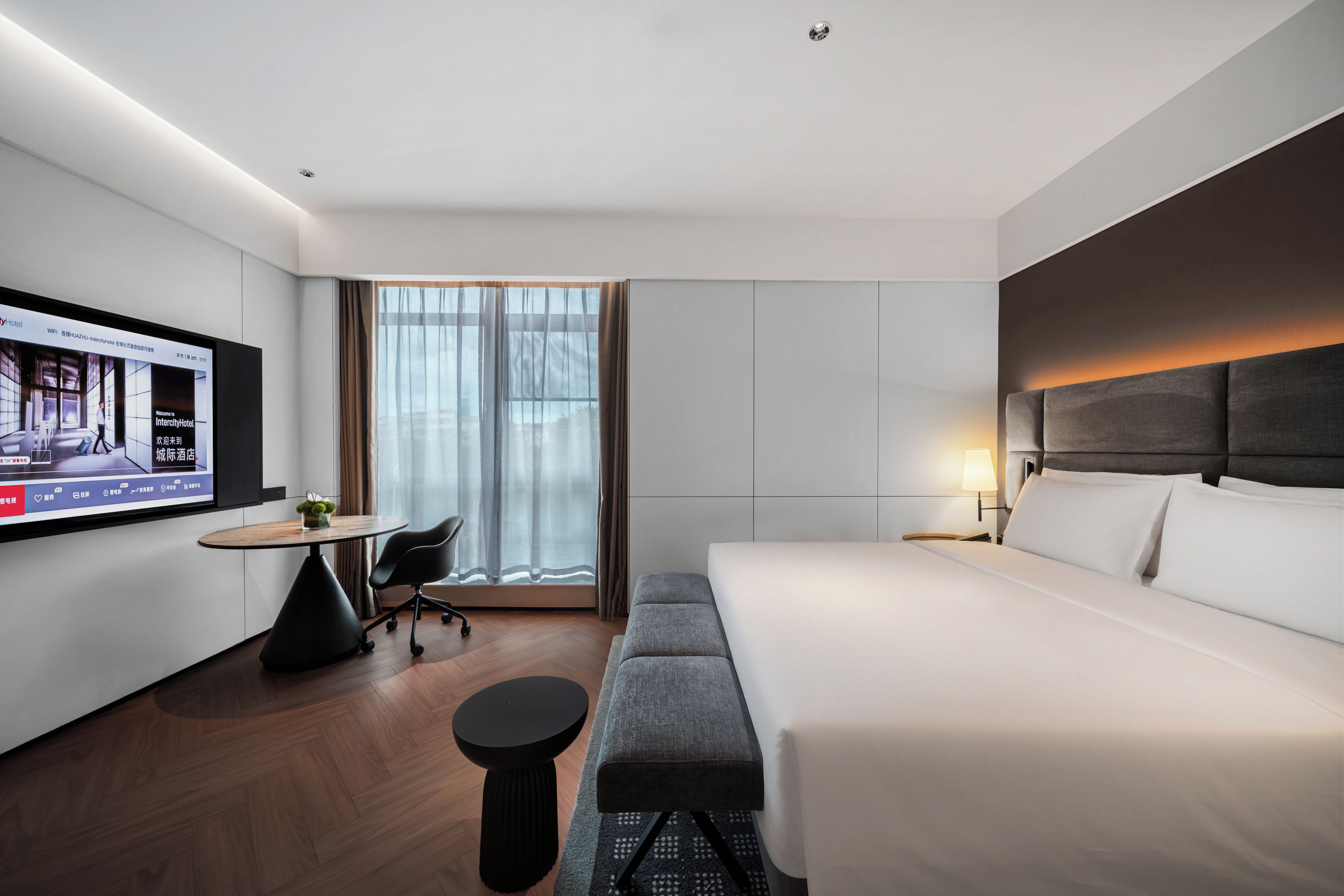 A modern hotel room with a large bed, a desk with a chair, a TV on the wall, and a window with curtains.