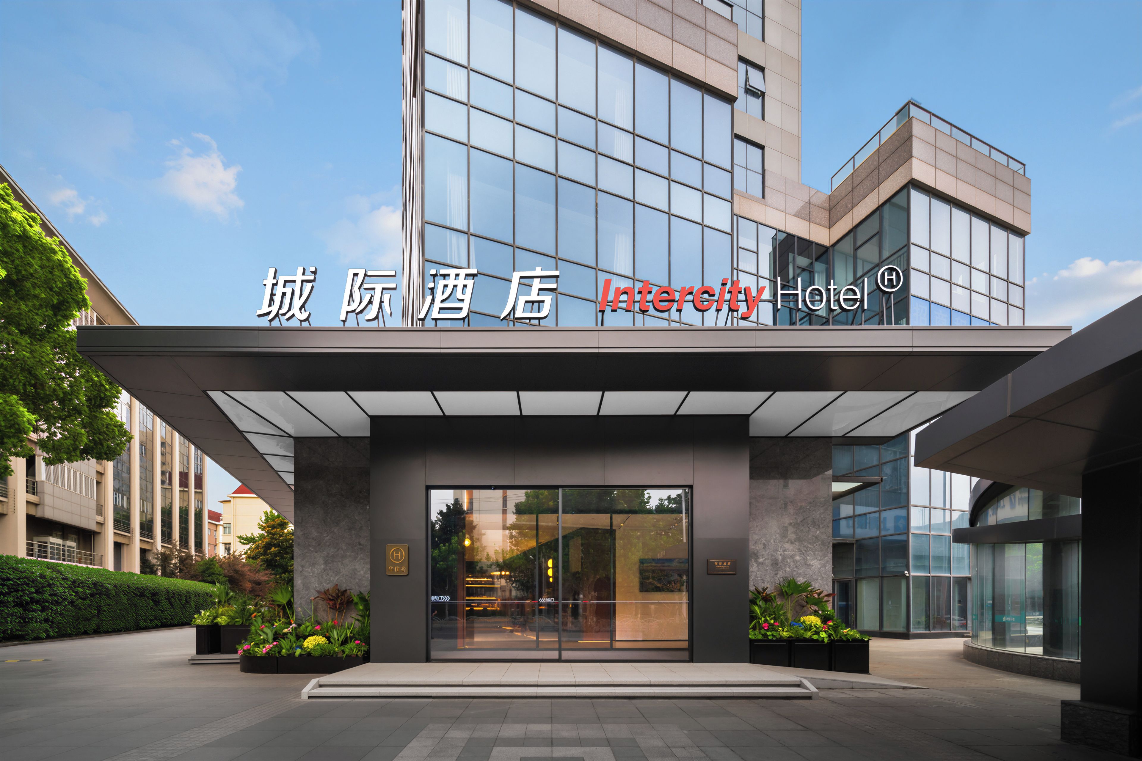 A modern hotel entrance with a glass facade, signage, and a landscaped area with planters.