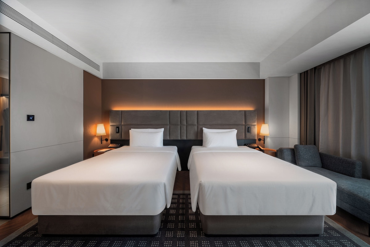 A hotel room with two beds, a sofa, a wall-mounted heater, and a digital thermostat.