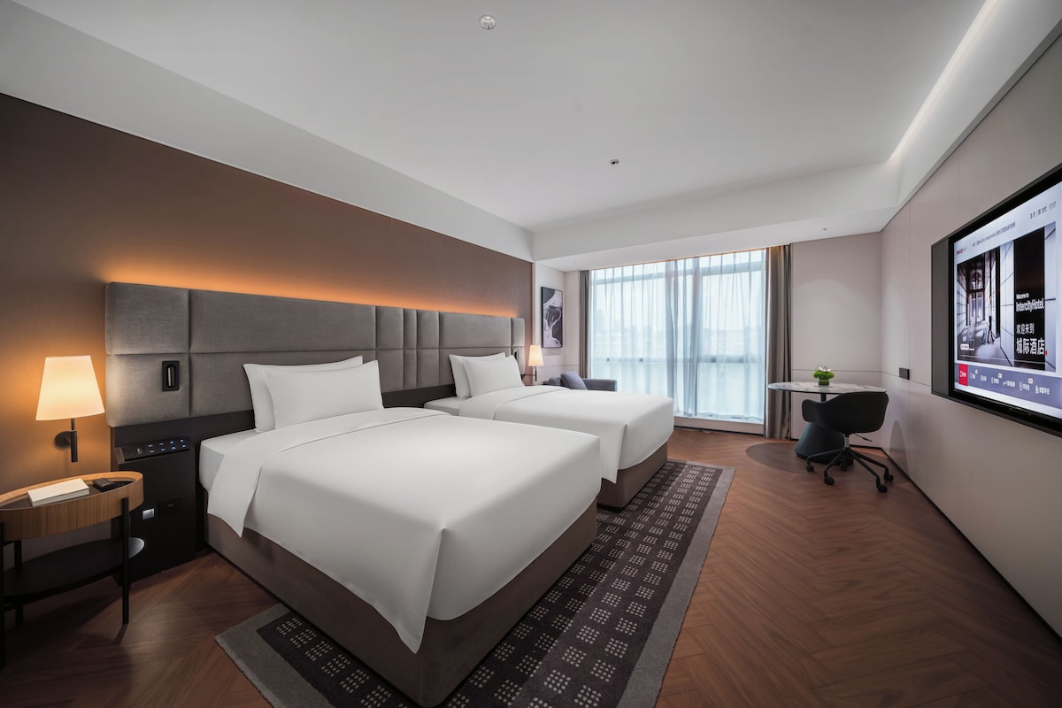 A hotel room with two beds, a desk, a chair, and a large window.