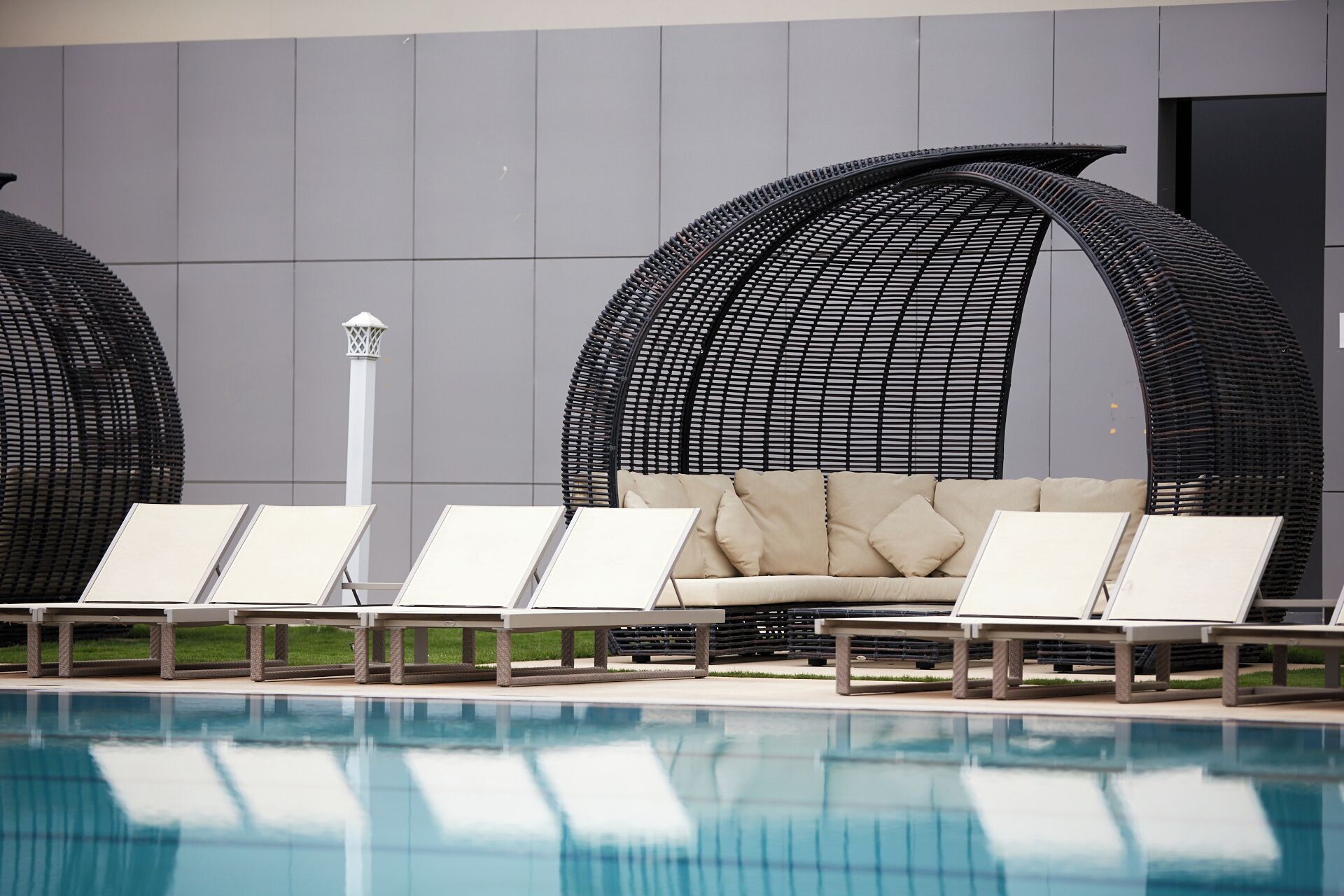 Un área junto a la piscina con sillones de descanso y un sofá, una piscina azul cristalina y la fachada moderna de un edificio.