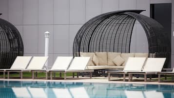 Un área junto a la piscina con sillones de descanso y un sofá, una piscina azul cristalina y la fachada moderna de un edificio.