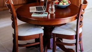 Un coin repas avec une table ronde en bois, quatre chaises, un ordinateur portable, un saladier de fruits et un verre d’eau.