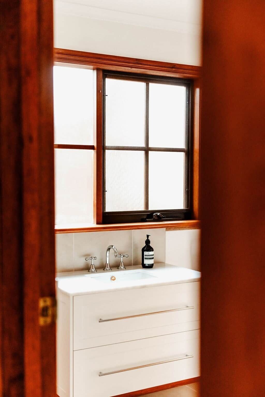 Un baño con un lavamanos blanco, una ventana encima y un marco de puerta de madera.