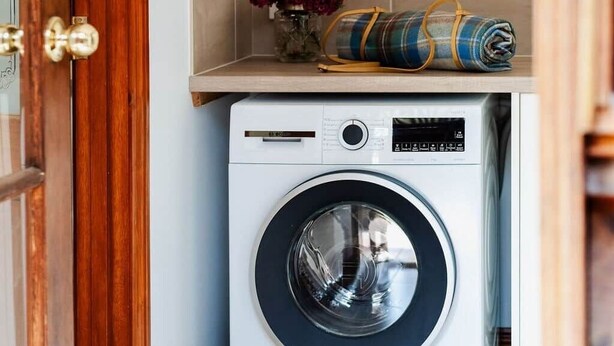 Une petite buanderie comprenant un lave-linge, une petite étagère ornée d’un vase de fleurs et une illustration botanique encadrée accrochée au mur.