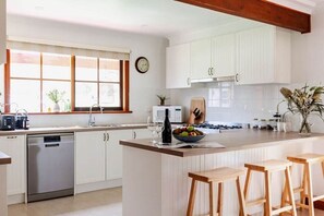 A kitchen with white cabinets, a built-in oven, a dishwasher, a window with blinds, and a wooden bar stool. - The Cottage at Paramoor Winery (Carlsruhe)