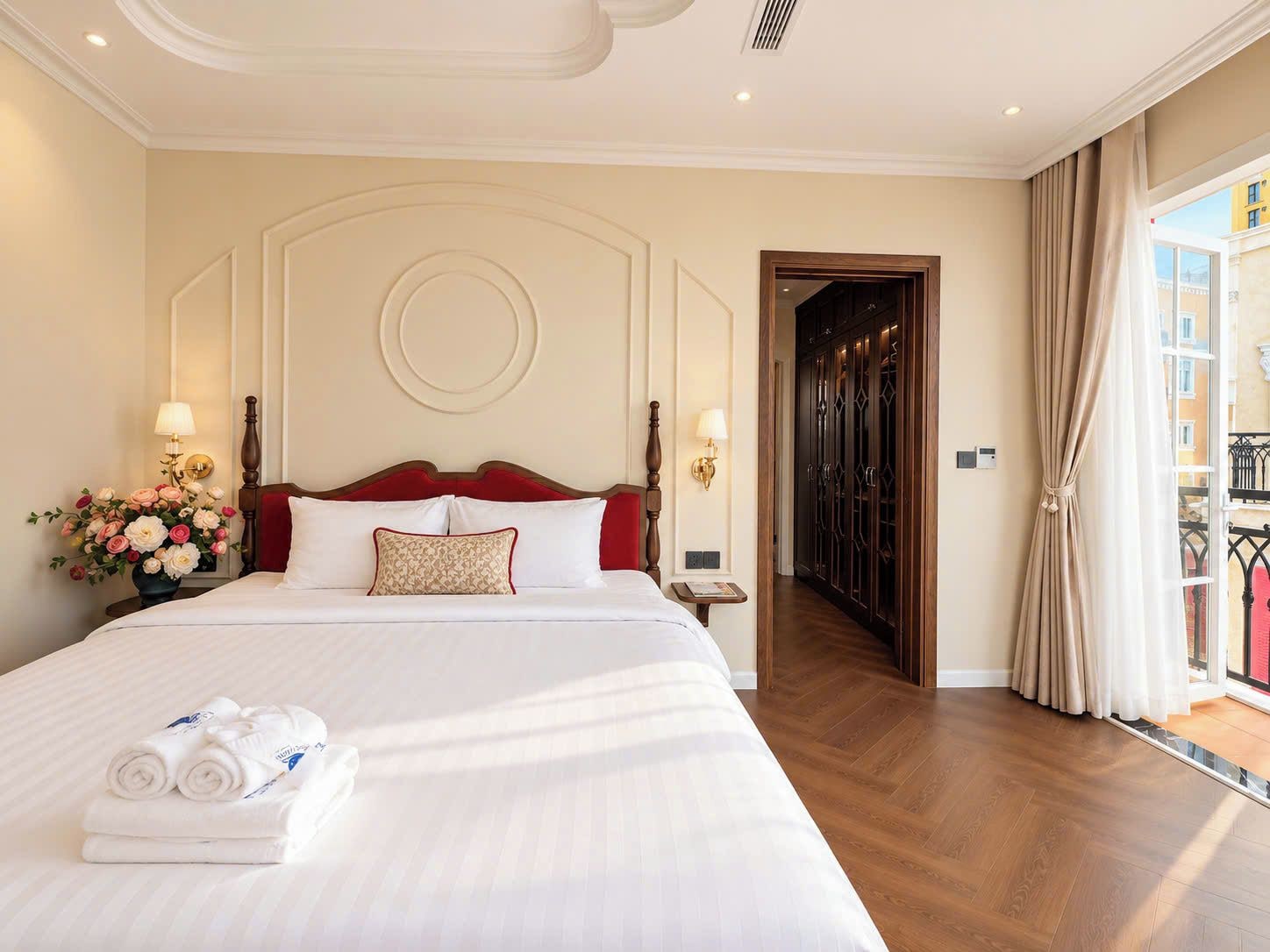 Habitación de hotel con una cama grande, una mesita de noche con lámpara, un jarrón de flores y vistas al exterior.