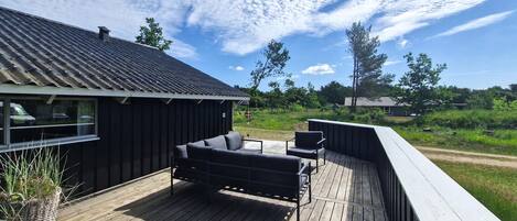 A wooden deck with outdoor furniture, a building with a metal roof, and a natural landscape in the background.