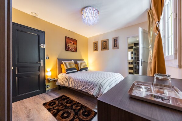 Un dormitorio con cama, lámparas de noche, mesita de noche, cómoda, espejo, ventana con cortinas y puerta.