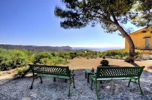 A view of a coastal landscape with a bench and table in the foreground, a yellow building to the right, and a clear sky.