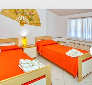 A room with two beds, a bedside table with a lamp, a window with curtains, and a decorative fan. - Apartment 'Aurora' with Sea View, Shared Pool and Wi-Fi (Poggi)