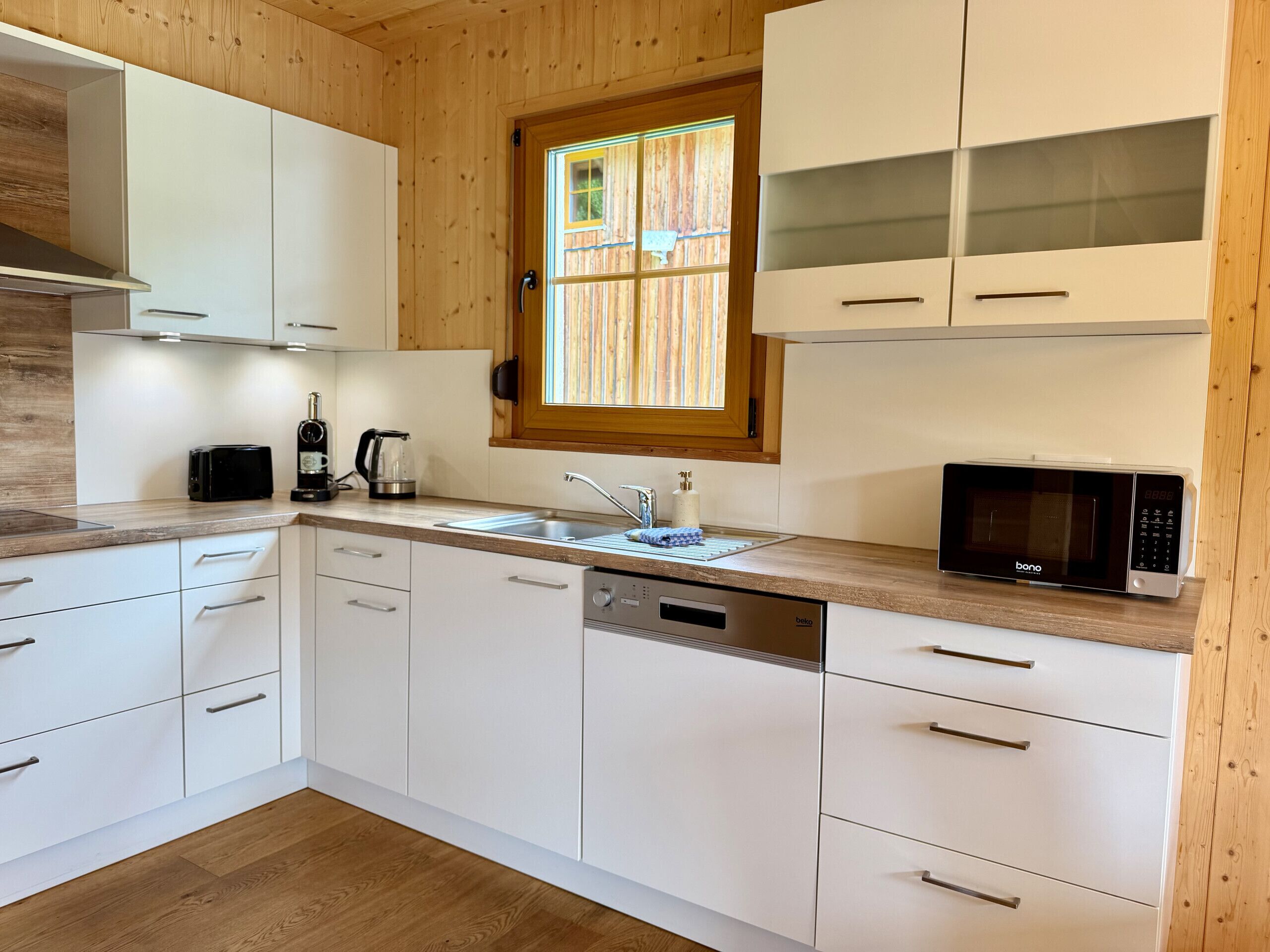 A modern kitchen with white cabinets, a built-in dishwasher, a microwave, and a window with blinds.