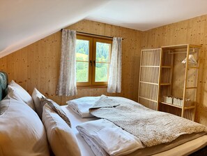 A bedroom with a wooden sloped ceiling, a large window with curtains, a bed with white linens, and a wooden wardrobe with hanging space.