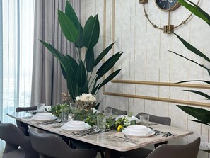 A dining table set with plates, glasses, and cutlery, surrounded by chairs, with a large plant and a wall-mounted clock. - Amazing Water View Luxury 2 Br Prime Location (Dubai)