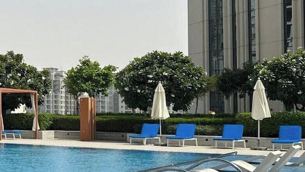 Une piscine entourée de chaises longues et de parasols, bordée de verdure, avec un grand immeuble en arrière-plan.