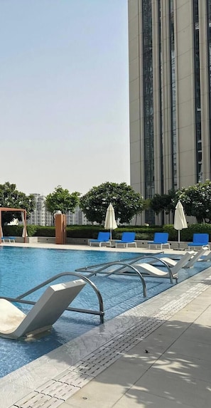 A swimming pool with lounge chairs and umbrellas, surrounded by greenery and a tall building in the background. - Amazing Water View Luxury 2 Br Prime Location (Dubai)